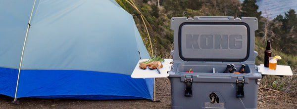 KONG 70 quart cooler open with two cutting boards attached. Cooler filled with ice and beverages next to a camping tent